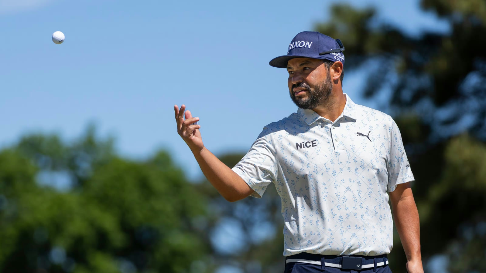 A tour pro tossing a golf ball on a sunny, tree-lined tournament course